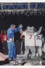 Two men hold a microphones on stage, a disc jockey, a grey dog mascot and other men stand in the background.