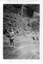 An unidentified little girl stands in the middle of a dirt road and is followed by a dog.
