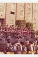 A group of men and women, wearing choir robes, stand on staging holding booklets at commencement.
