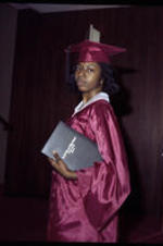 An unidentified young woman stands holding her diploma under her arm.