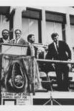 Ralph D. Abernathy is shown speaking at a Poor People's Campaign event in Memphis, Tennessee. To Abernathy's right on the stage are Senator Ted Kennedy, Evelyn G. Lowery, and Juanita Abernathy. A cut-out photo of Joseph E. Lowery from the 1980s is pasted onto the photo next to Abernathy.