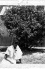 An unidentified woman in white dress sits outside on the lawn.