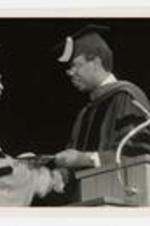 President Elias Blake Jr. shakes hands with another man, wearing graduation caps and gowns, on stage at commencement.