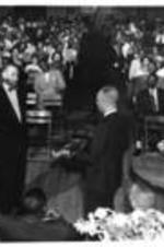 John H. Wheeler stands with an unidentified man in front of a crowd while another man presents a plaque.