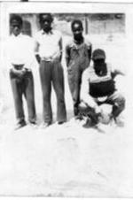 Three boys stand next to a crouching baseball catcher.