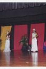 Four women, wearing matching floor-length silver dressed with tiaras and sashes, hold bouquets of flowers on stage.